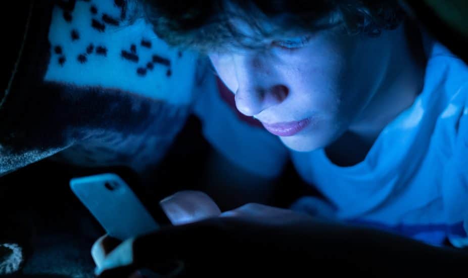 young boy using a smartphone lying in bed under blanket at night