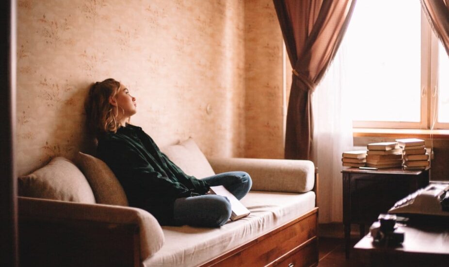 Young upset woman holding book looking through window while sitting on sofa in living room at home